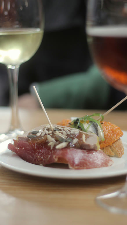 Un pintxo servido en un plato, con dos copas detrás